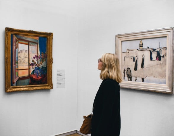 Blonde woman in glasses observes a framed abstract artwork on a white gallery wall. Another landscape painting and a text plaque are on an adjacent wall.