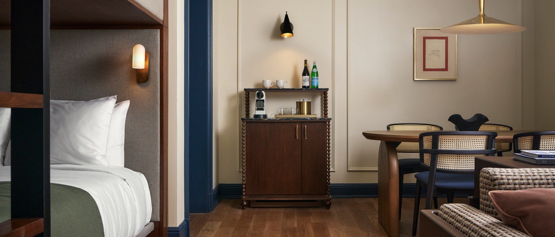 A hotel room features a bunk bed, a minibar cabinet, and a dining table with chairs. Bottles of PELLEGRINO are on the cabinet; a "DOORWAY" book is on a sofa.