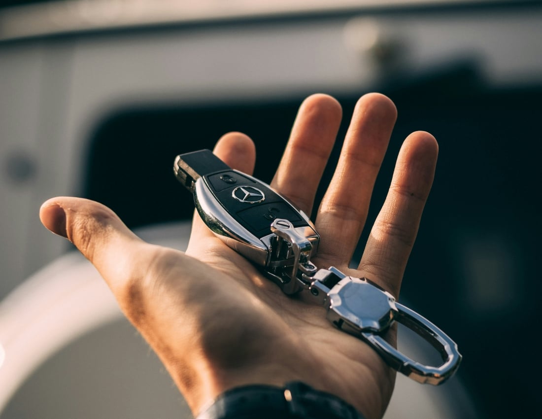 A hand holds a black and silver Mercedes-Benz car key and metallic keychain, palm up, in front of a blurred white vehicle outdoors.