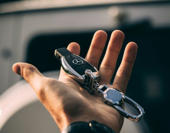 A hand holds a black and silver Mercedes-Benz car key and metallic keychain, palm up, in front of a blurred white vehicle outdoors.