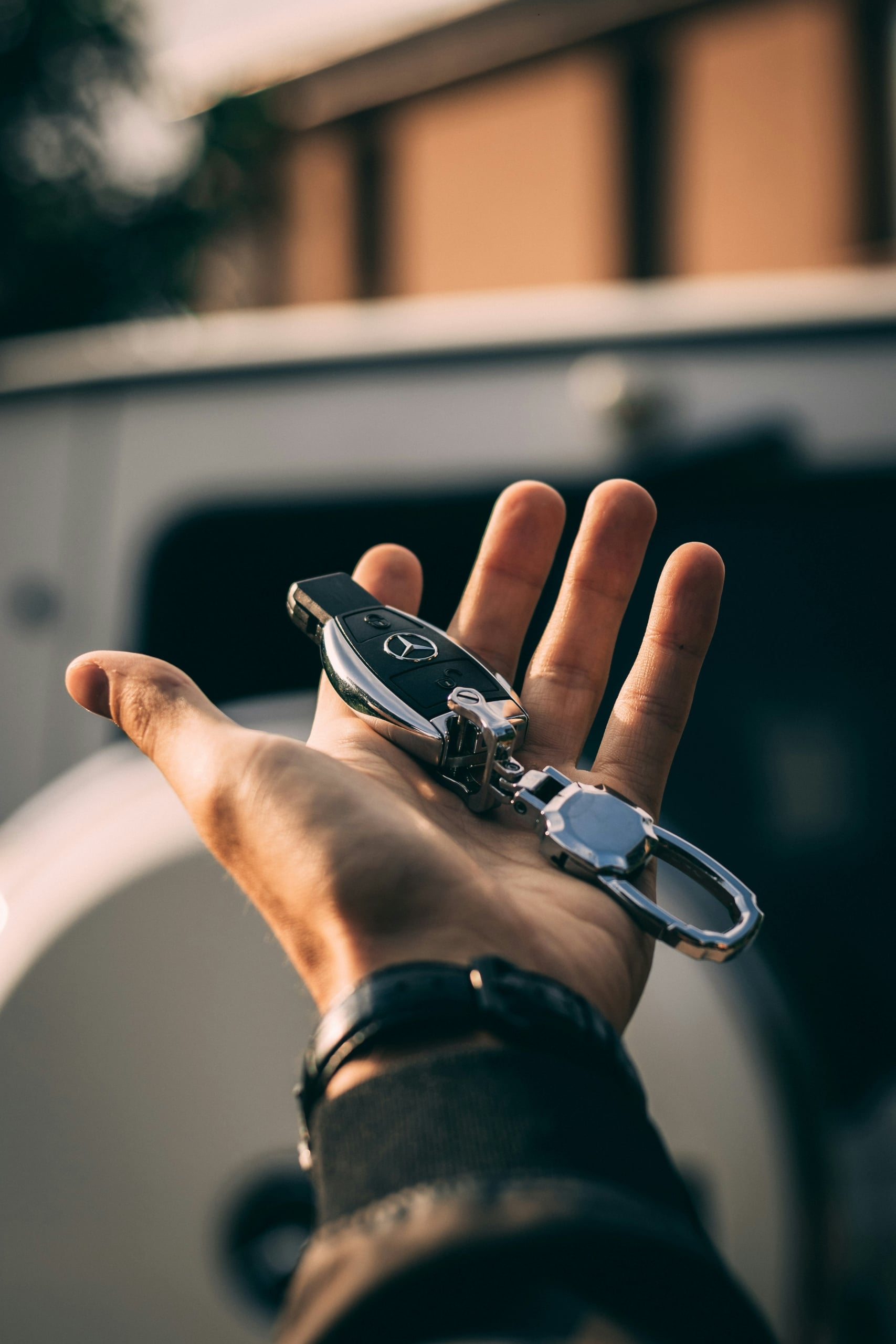 A hand holds a black and silver Mercedes-Benz car key and metallic keychain, palm up, in front of a blurred white vehicle outdoors.