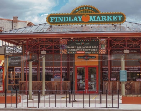 Findlay Market building stands under a cloudy sky. Its entrance features a sign and banners, with a protective railing in front.