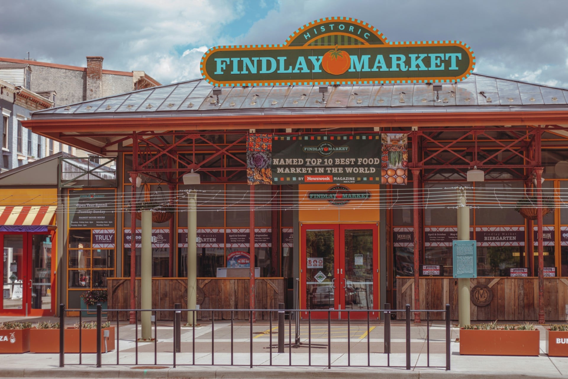 Findlay Market building stands under a cloudy sky. Its entrance features a sign and banners, with a protective railing in front.