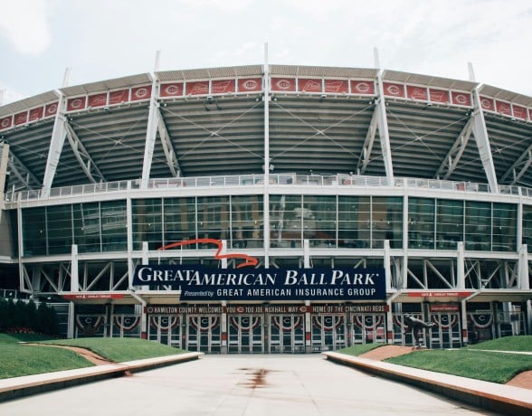Front view of a large, multi-tiered baseball stadium featuring its main entrance from a broad plaza with landscaped grassy areas, under a light sky.