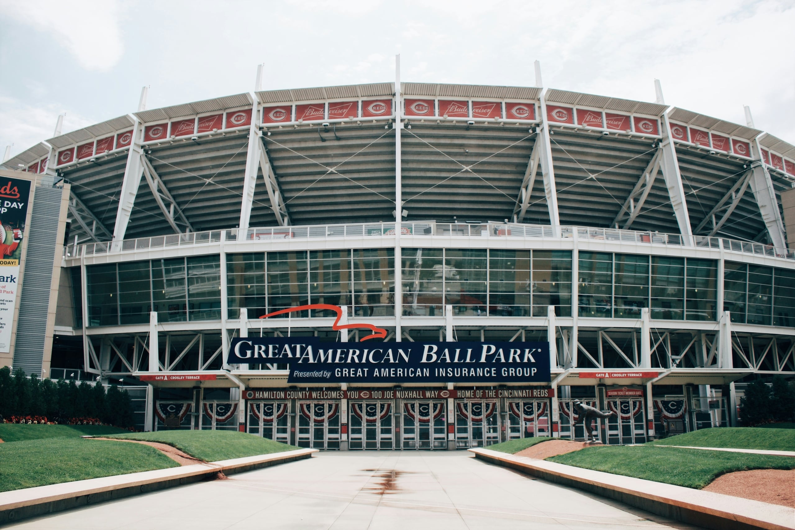 Front view of a large, multi-tiered baseball stadium featuring its main entrance from a broad plaza with landscaped grassy areas, under a light sky.