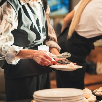 A person in a black vest and patterned shirt uses tongs to serve a pastry onto a white plate. They are at a dessert buffet with various sweets and stacked plates, surrounded by other people.