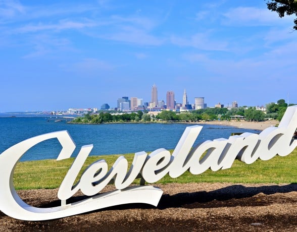 Large white "Cleveland" letters stand on a grassy hill overlooking a blue lake with a distant city skyline and sandy beach under a clear sky.
