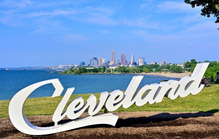 Large white "Cleveland" letters stand on a grassy hill overlooking a blue lake with a distant city skyline and sandy beach under a clear sky.