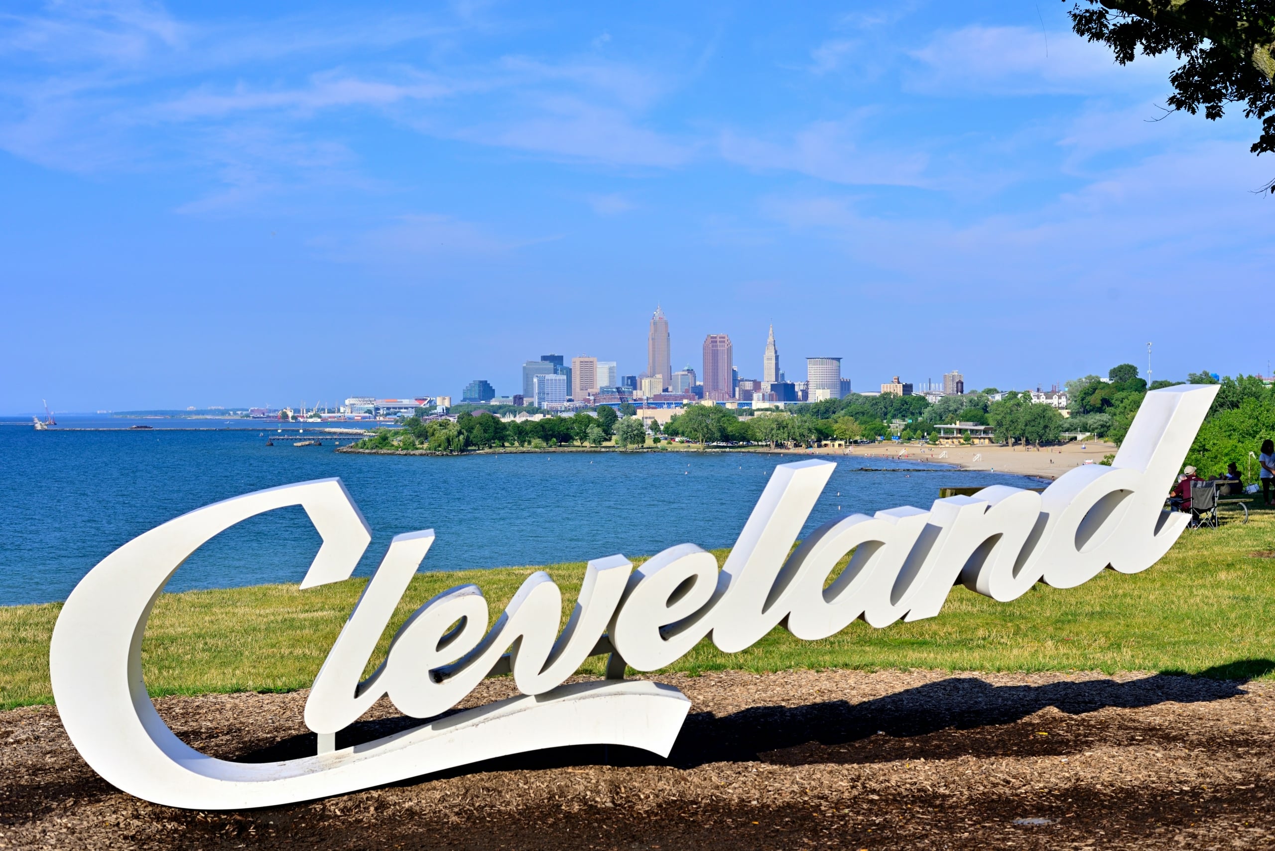 Large white "Cleveland" letters stand on a grassy hill overlooking a blue lake with a distant city skyline and sandy beach under a clear sky.