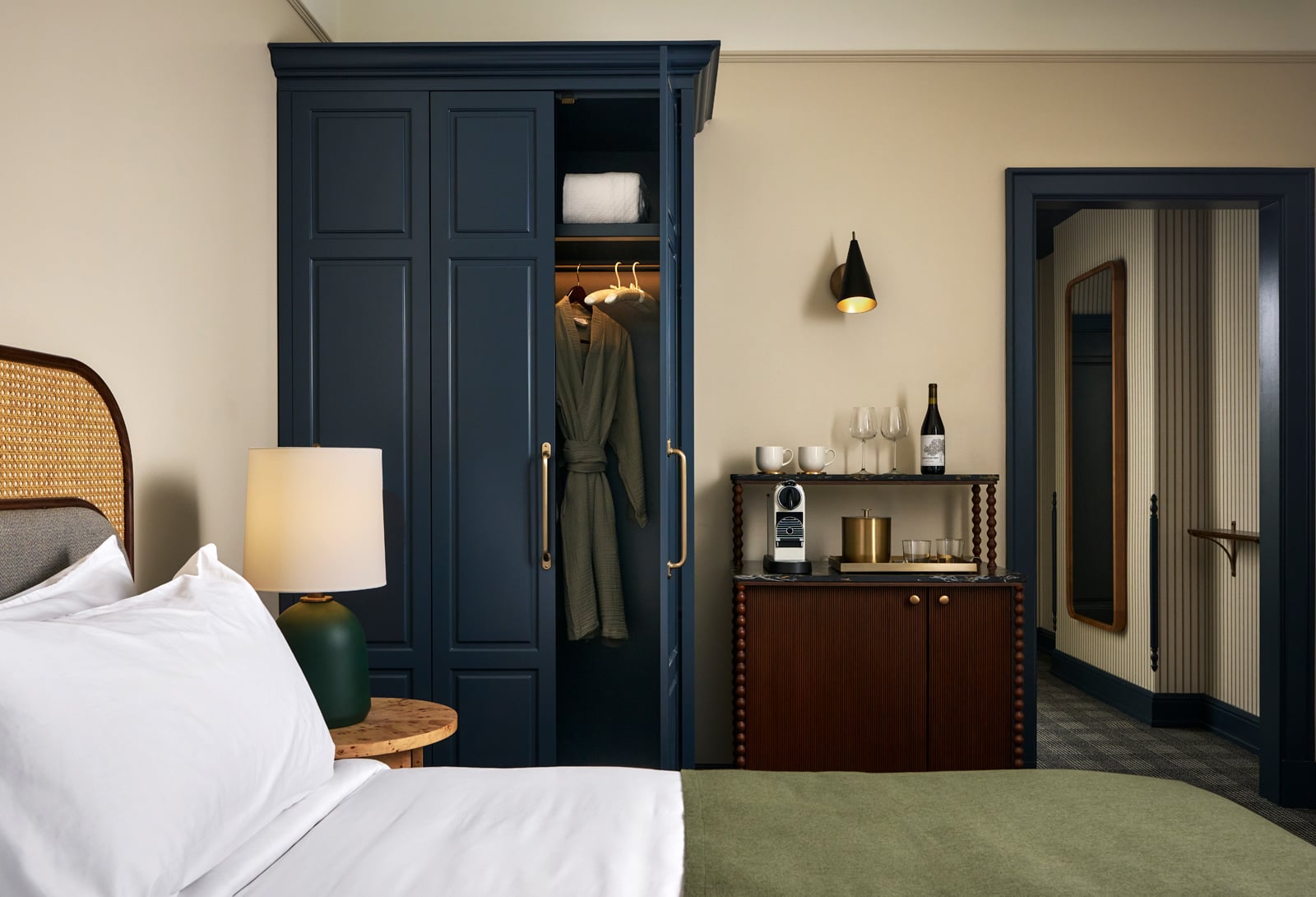 A dark blue wardrobe stands with its right door ajar, revealing a green bathrobe and towel. A mini-bar cabinet displays a coffee maker, wine, and glasses in a modern hotel room.