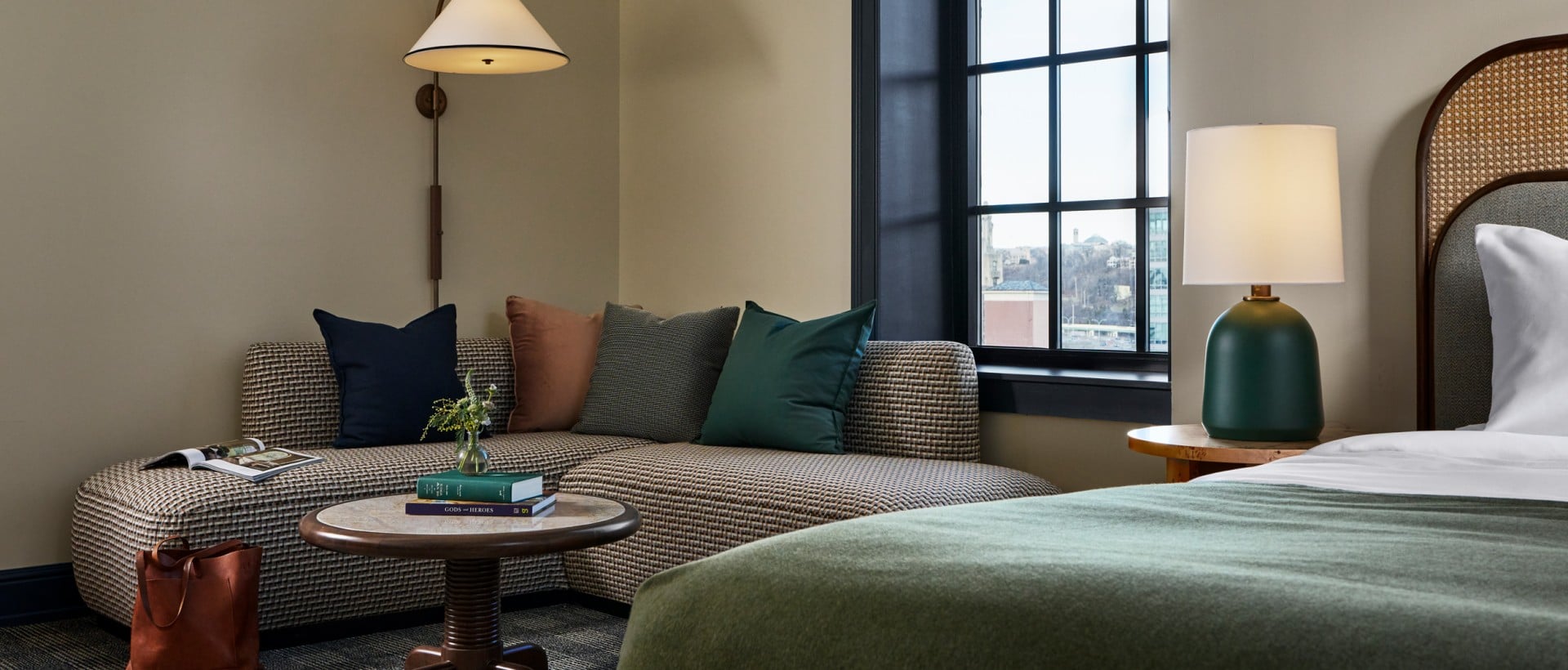 A patterned corner sofa with colorful pillows and a round coffee table occupies a modern room corner. A wall lamp, large grid window, and bed are visible. Books: GODS and HEROES, ART and ARCHITECTURE.