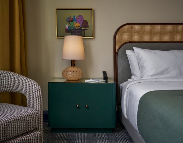 Green bedside table with a textured lamp, notebook, and phone stand, sits next to a bed with a woven headboard. A patterned armchair and yellow curtain are visible.