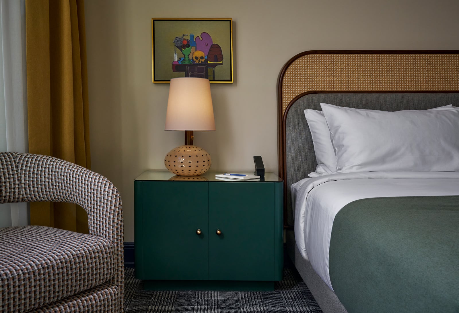 Green bedside table with a textured lamp, notebook, and phone stand, sits next to a bed with a woven headboard. A patterned armchair and yellow curtain are visible.