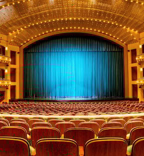 A grand theater auditorium with rows of empty red seats faces a large stage. A closed dark blue curtain is brightly illuminated by blue light under ornate, lit ceilings.