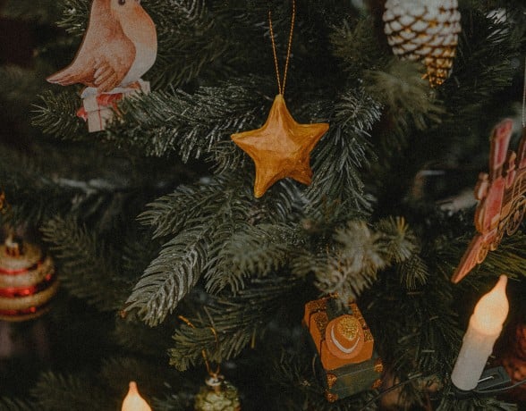 A Christmas tree displays various ornaments: a bird in a hat, a golden star, pinecones, and a tank. Warm candle lights glow amidst the dark green branches.