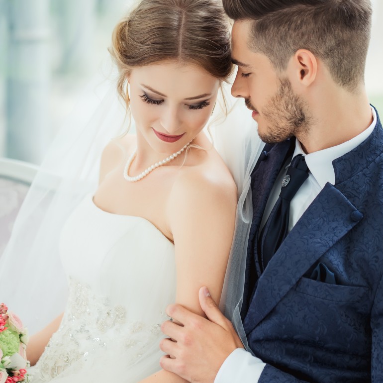 A bride in a white gown and pearl necklace holds a bouquet, looking down while the groom in a blue patterned suit embraces her, foreheads touching, seated indoors.