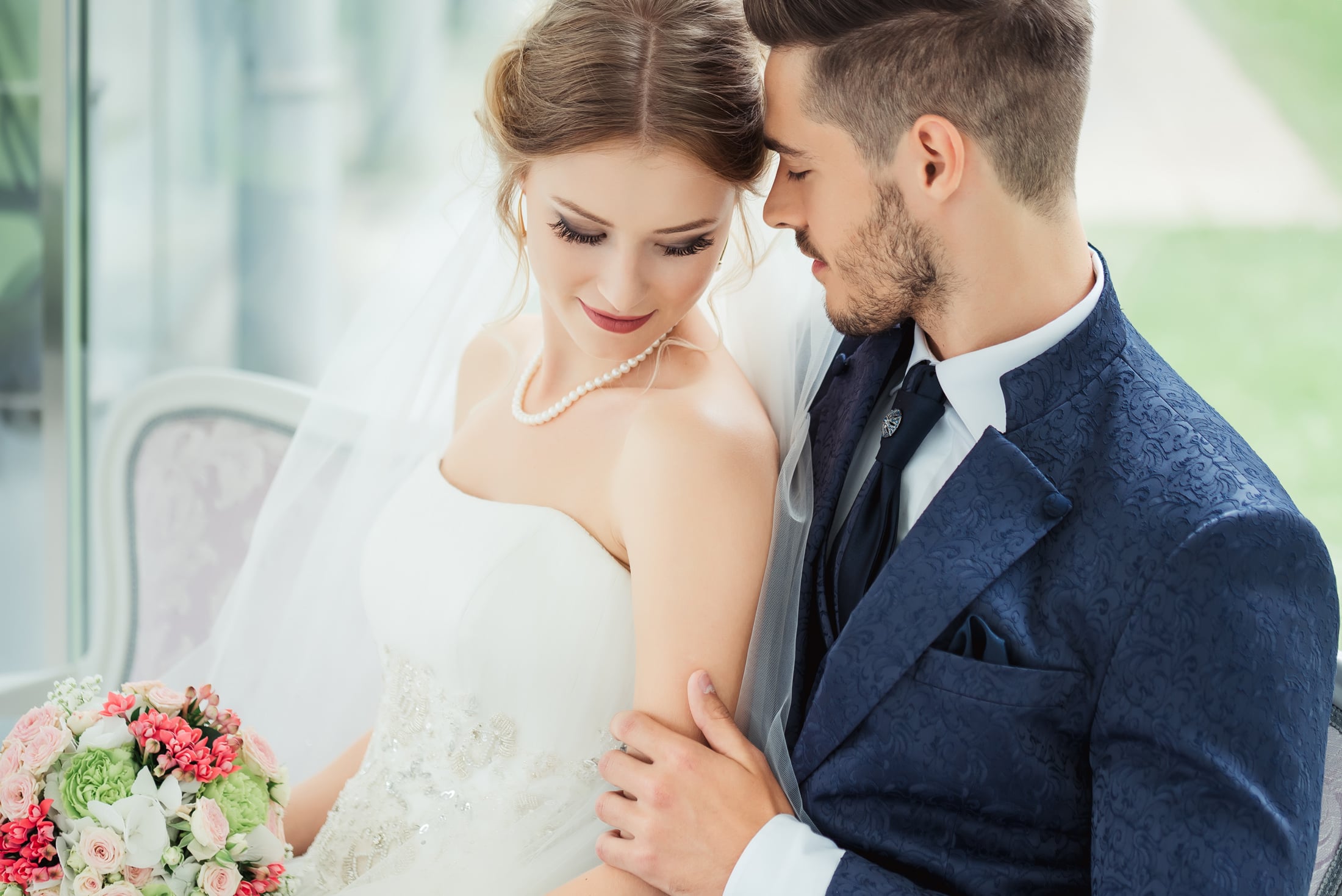 A bride in a white gown and pearl necklace holds a bouquet, looking down while the groom in a blue patterned suit embraces her, foreheads touching, seated indoors.