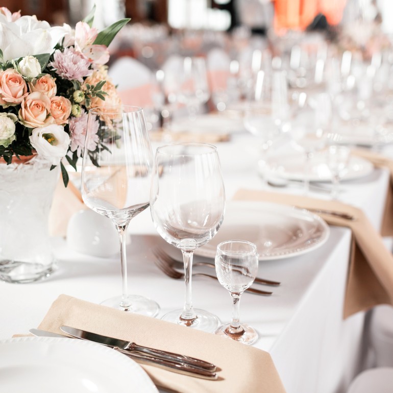 A vibrant floral centerpiece adorns a formal dining table, surrounded by wine glasses, plates, and cutlery on a white tablecloth, set for an event.