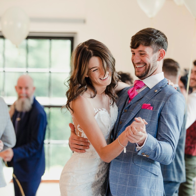 A smiling bride in a white dress and groom in a blue suit dance joyfully, embracing each other. They are surrounded by other guests and white balloons at a reception.