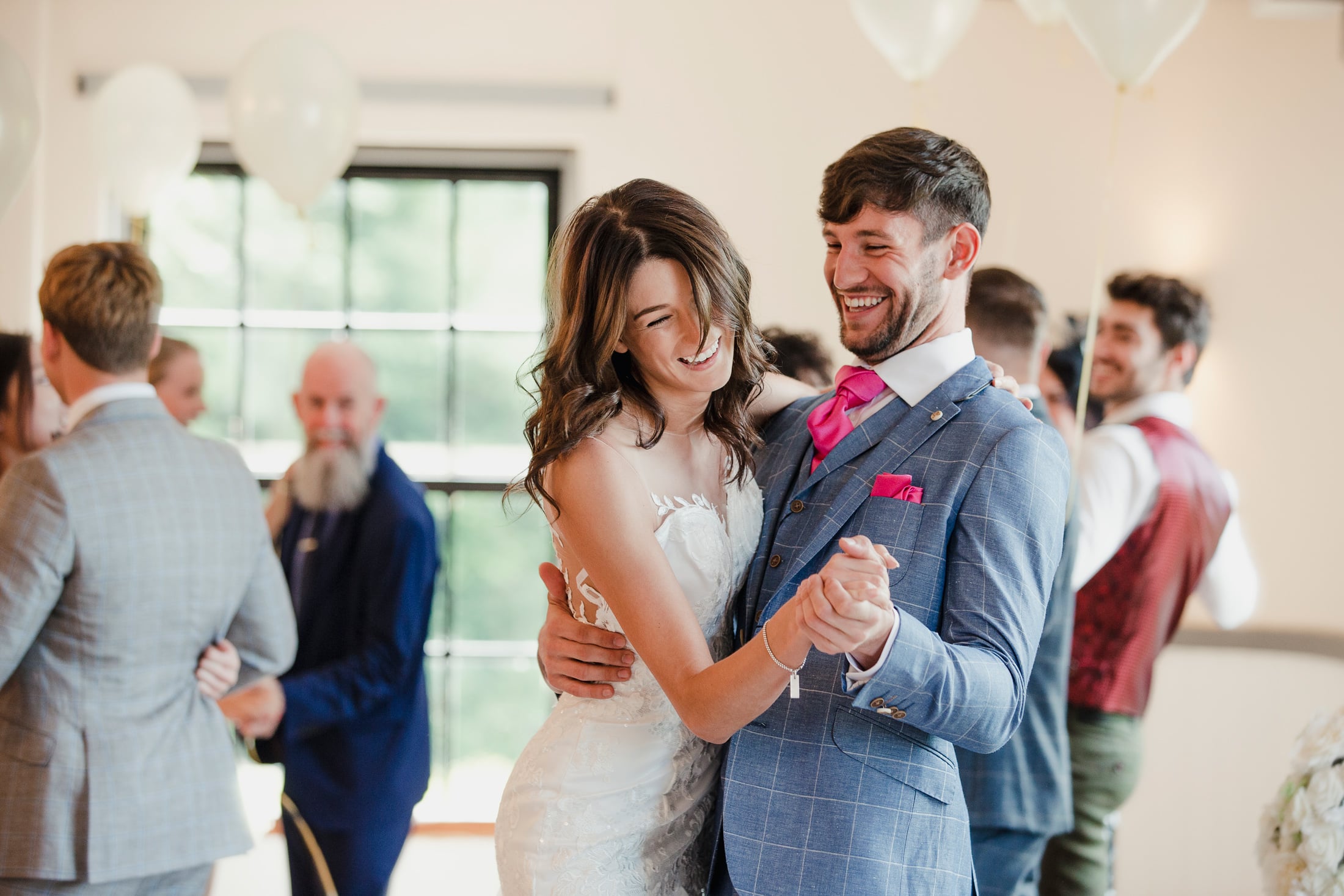 A smiling bride in a white dress and groom in a blue suit dance joyfully, embracing each other. They are surrounded by other guests and white balloons at a reception.