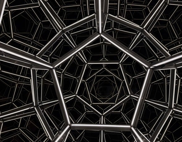 Geometric metal framework forms repeating pentagonal and hexagonal shapes against a black background, creating a tunnel-like visual effect.