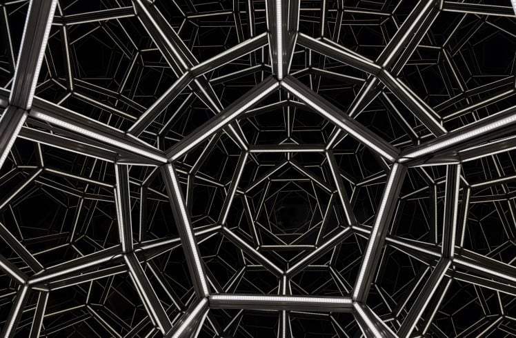 Geometric metal framework forms repeating pentagonal and hexagonal shapes against a black background, creating a tunnel-like visual effect.