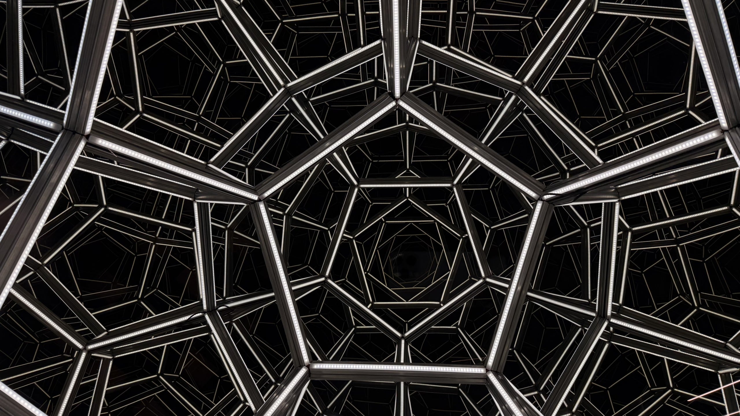 Geometric metal framework forms repeating pentagonal and hexagonal shapes against a black background, creating a tunnel-like visual effect.
