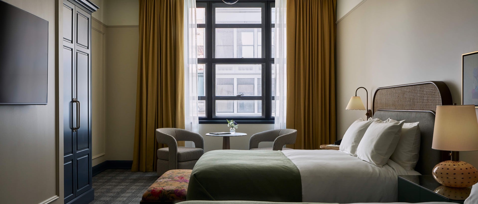 Hotel Room with Two Queen Beds on right side of wall, TV mounted on left side, with a large window surrounded by gold panel curtains.