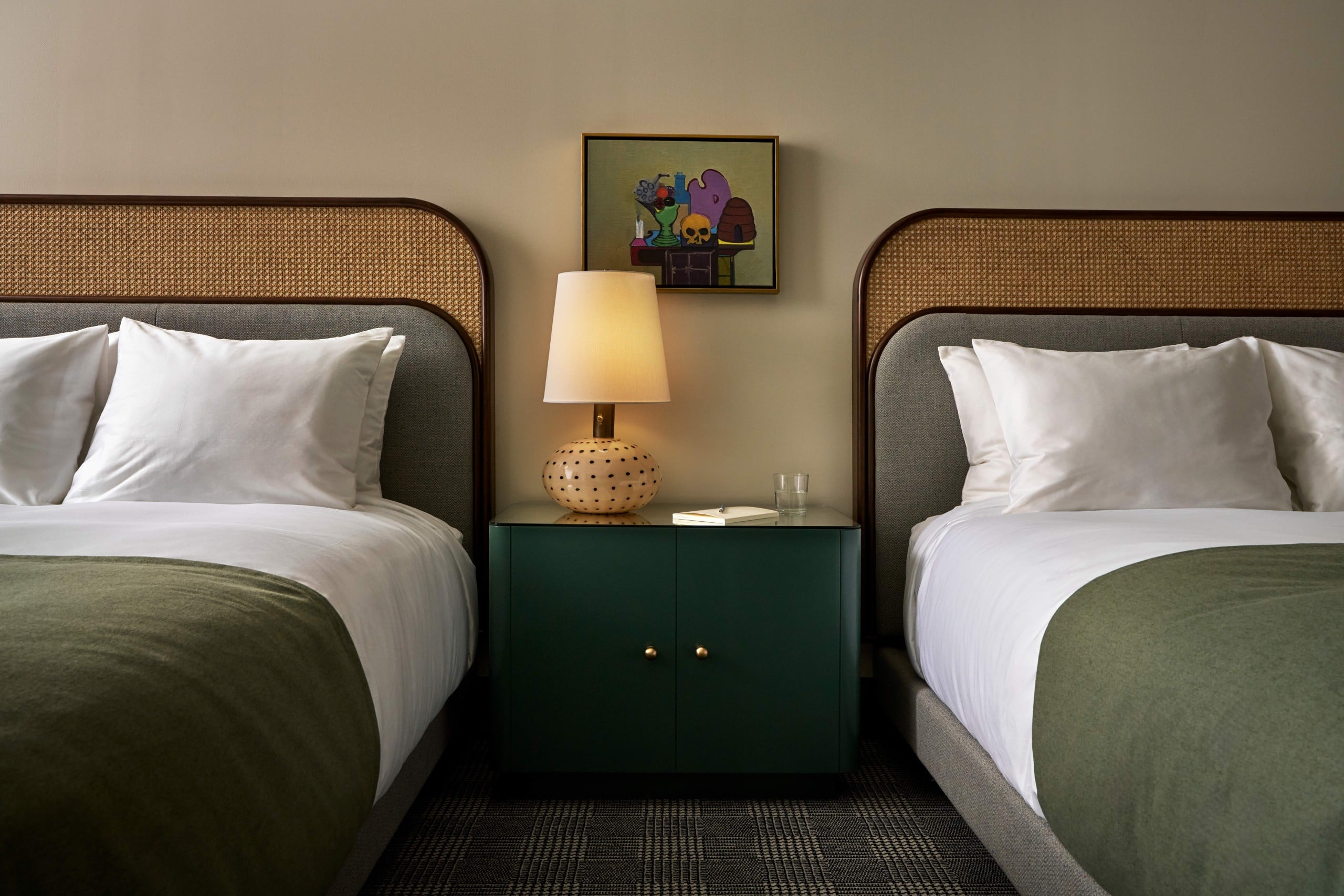 Two queen sized beds with white linen and green blanket with nightstand in middle topped with a lamp and artwork in the background.