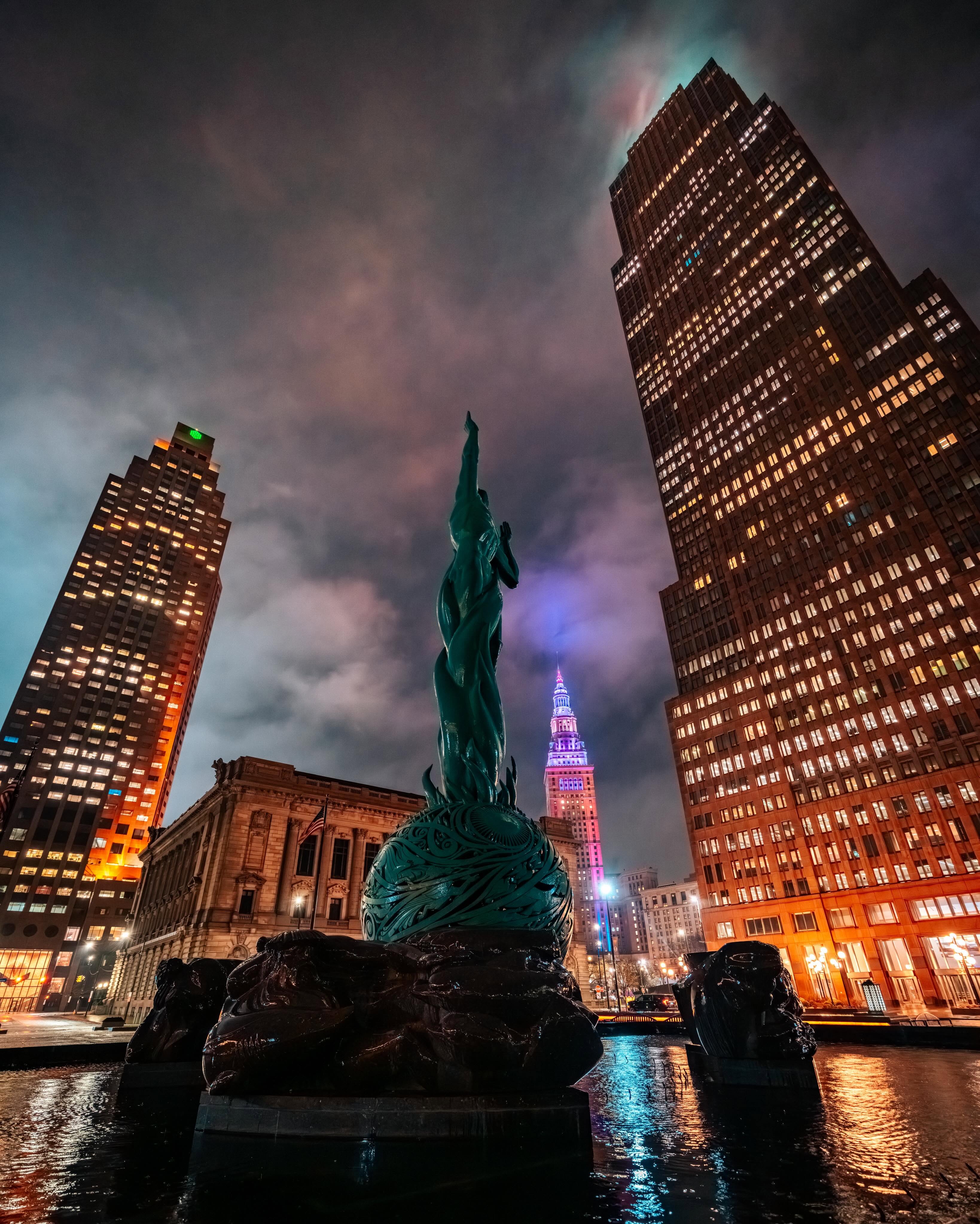 A nighttime cityscape with a prominent statue in the foreground and skyscrapers in the background.